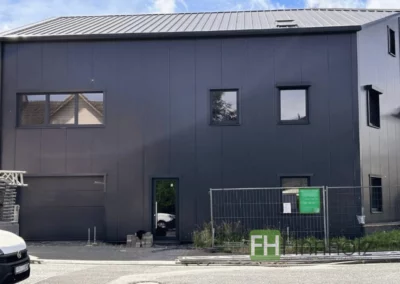 Wohnungsbau in Frankfurt am Main - Bild 2: Das Bild zeigt erneut eine Außenansicht der mehrstöckigen Wohnung mit schwarzer Fassade. Diesmal in der Frontalansicht. Auch hier ist das trapezförmige Dach sehr gut zu erkennen.